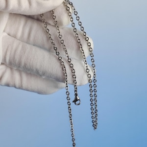 May include: A silver-coloured necklace with a delicate chain design. The necklace is held by a gloved hand against a light blue background. The chain has a lobster clasp closure.