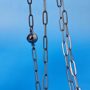 Peut inclure: Gros plan sur un collier chaîne gris foncé avec un design à maillons ovales. Une sphère brillante de couleur argentée est suspendue à la chaîne. Le fond est de couleur bleu vif uni.