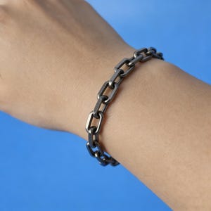 Könnte beinhalten: Ein dunkelgraues Kettenarmband mit rechteckigen Gliedern wird an einem Handgelenk vor blauem Hintergrund getragen. Das Armband besteht aus Metall und hat ein schlichtes, industrielles Design. Die Glieder sind gleichmäßig angeordnet und erzeugen einen kühnen, modernen Look.
