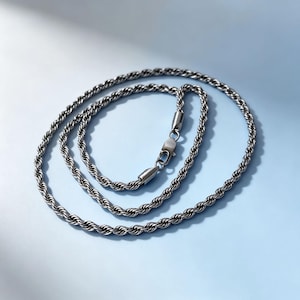 May include: A dark silver-toned rope chain necklace featuring a twisted metal link design. The necklace is arranged in a circular coil and includes a secure lobster claw clasp mechanism at the center, connecting the two ends of the chain.
