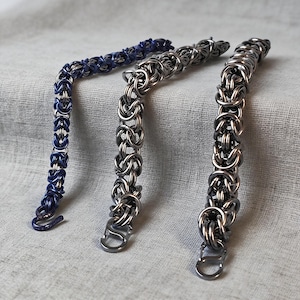 May include: Three chainmail bracelets featuring a complex Byzantine weave pattern. One bracelet is a deep metallic blue, while the other two are a polished silver-tone metal. Each bracelet terminates in a simple S-shaped hook clasp for closure.