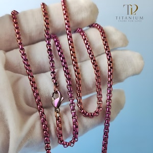 May include: A pink and gold-toned necklace with a lobster clasp. The chain is made of small, connected links. The necklace is displayed on a white surface. The logo "TITANIUM STORE FOR LOVE" is in the upper right corner.