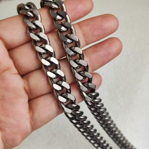 May include: Two silver curb chains, one thicker than the other, are shown laid out on a hand. The chains are made of metal and have a shiny finish.