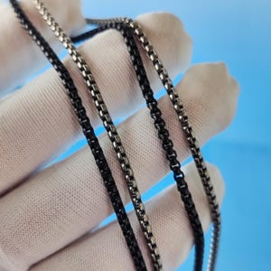 May include: Close-up of several metal chain necklaces in varying shades of black and silver. The chains are made of small, square links, creating a textured appearance. The background is a light blue.