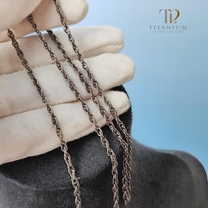 以下が含まれることがあります： シルバーカラーのチェーンネックレスのクローズアップ。チェーンは白い手袋と青い背景に表示されています。ネックレスはねじれたロープのデザインです。画像には「TITANIUM STORE FOR LOVE」というテキストも含まれています。