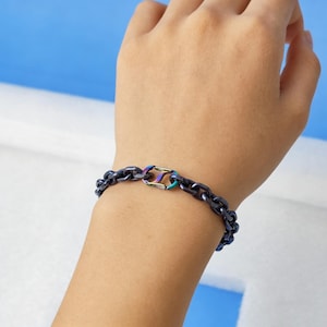 Könnte beinhalten: Ein dunkelblaues Kettenarmband mit einem regenbogenfarbenen Verschluss. Das Armband besteht aus verbundenen Metallteilen und wird am Handgelenk getragen. Der Hintergrund ist hellblau und weiß.