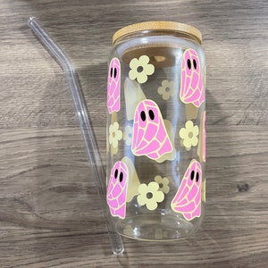 May include: A clear glass can-shaped tumbler with a bamboo lid and a clear glass straw. The tumbler is decorated with pink ghost and yellow flower designs. The tumbler is designed to hold approximately 16 ounces.