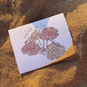 May include: A white greeting card with a botanical illustration of a monstera plant in shades of pink and beige. The card is on a sandy surface, and the text "mahalo nui loa" is written in cursive.