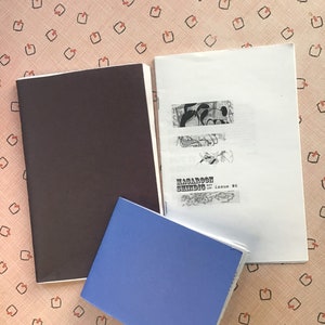 Op de afbeelding: Drie zines worden getoond op een oppervlak met een patroon. E&eacute;n is een gesloten, donkerbruin boekje. Een andere is open, met zwart-wit illustraties en de tekst "MACAROON SHINDIC issue #6". Een blauw boekje heeft de tekst "Germoon Shandig #4".