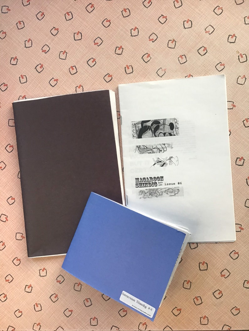 Op de afbeelding: Drie zines worden getoond op een oppervlak met een patroon. E&eacute;n is een gesloten, donkerbruin boekje. Een andere is open, met zwart-wit illustraties en de tekst "MACAROON SHINDIC issue #6". Een blauw boekje heeft de tekst "Germoon Shandig #4".