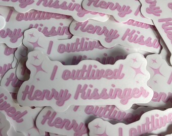 Pode incluir: Uma pilha de adesivos rosa e branco com o texto "I outlived Henry Kissinger" em uma fonte rosa cursiva. Os adesivos têm um design brilhante.