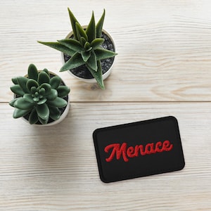 May include: A black rectangular patch with the word "Menace" embroidered in red cursive lettering. The patch is on a white wooden surface with two small potted green plants.