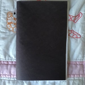 Op de afbeelding: Een gesloten, rechthoekig notitieboek met een donkerbruine kaft. Het notitieboek is verticaal geplaatst, met de rug aan de linkerkant. De achtergrond is een lichtblauwe en witte quilt met geborduurde details.