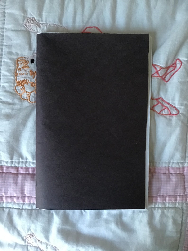 Op de afbeelding: Een gesloten, rechthoekig notitieboek met een donkerbruine kaft. Het notitieboek is verticaal geplaatst, met de rug aan de linkerkant. De achtergrond is een lichtblauwe en witte quilt met geborduurde details.