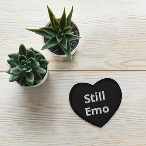 May include: A black heart-shaped patch with the words "Still Emo" embroidered in white. Two small potted succulents are also visible, one with rounded leaves and the other with pointed leaves. The background is a light wood grain.