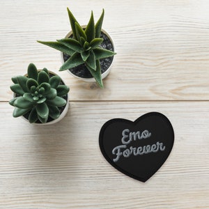 May include: A black heart-shaped patch with the words "Emo Forever" embroidered in silver thread. Two small potted succulents sit on a light wood-grain surface.