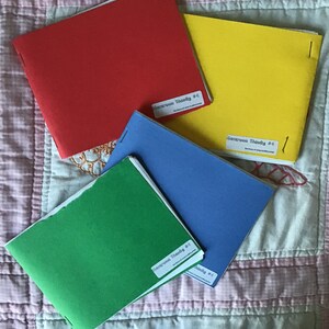 Op de afbeelding: Vier kleurrijke boekjes: rood, geel, blauw en groen. Elk boekje heeft een wit etiket met zwarte tekst: "Kierkegaard Shards #4 The House of Many Buddhist Press". De boekjes liggen op een roze en witte quilt.