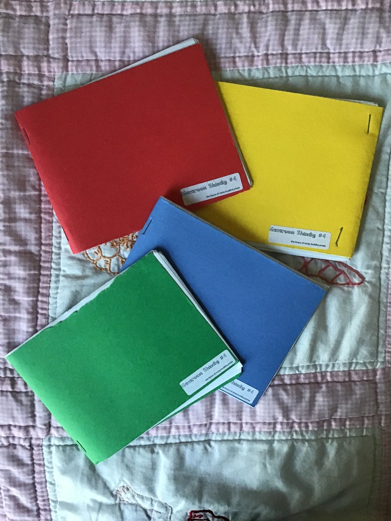 Op de afbeelding: Vier kleurrijke boekjes: rood, geel, blauw en groen. Elk boekje heeft een wit etiket met zwarte tekst: "Kierkegaard Shards #4 The House of Many Buddhist Press". De boekjes liggen op een roze en witte quilt.