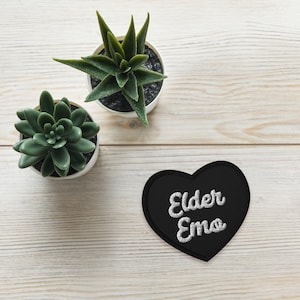 May include: A black heart-shaped patch with the words "Elder Emo" embroidered in white. Two small potted succulents sit on a light wood-grain surface.