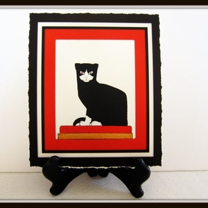 May include: A black and white cat with a red and gold mat sits in a red frame with a black mat. The image is framed and on a black stand.