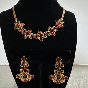 May include: A gold necklace and earrings set with a delicate design featuring red gemstones. The necklace is a chain with a pendant of interconnected swirls and the earrings are dangling with a similar design.