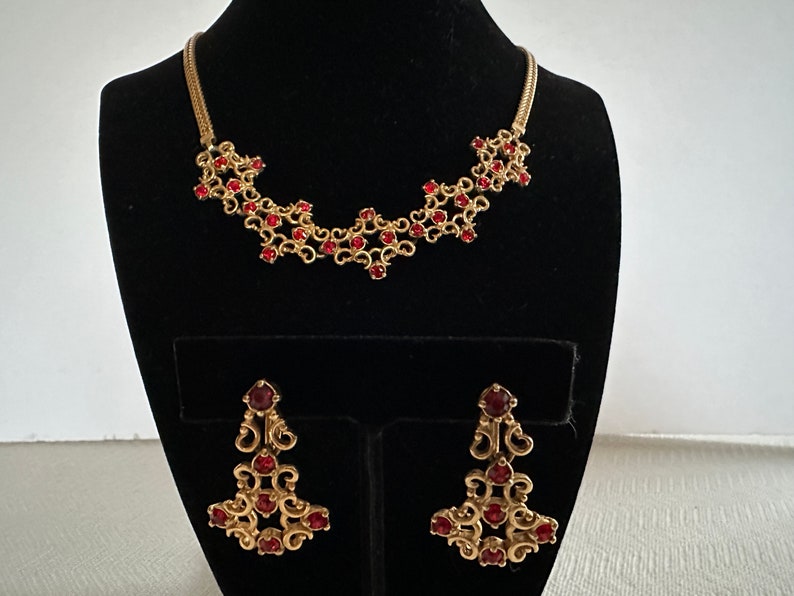 May include: A gold necklace and earrings set with a delicate design featuring red gemstones. The necklace is a chain with a pendant of interconnected swirls and the earrings are dangling with a similar design.