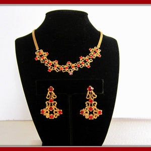May include: A gold necklace and earrings set with red rhinestone accents. The necklace is a delicate chain with a floral design and the earrings are dangling with a similar floral design.