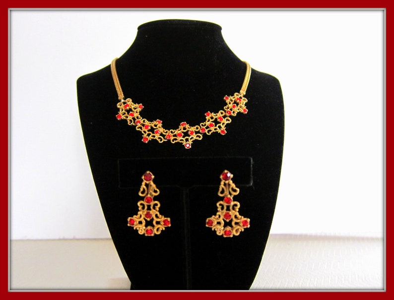 May include: A gold necklace and earrings set with red rhinestone accents. The necklace is a delicate chain with a floral design and the earrings are dangling with a similar floral design.