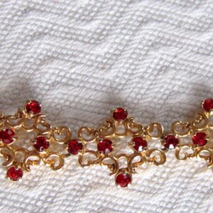 May include: A gold necklace with a delicate, intricate design featuring a repeating pattern of small red gemstones.