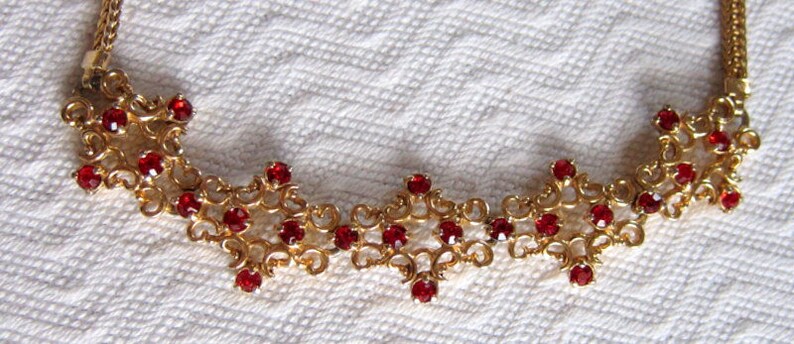 May include: A gold necklace with a delicate, intricate design featuring a repeating pattern of small red gemstones.