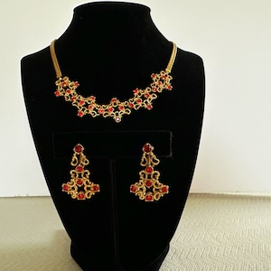 May include: A gold necklace and earrings set with red rhinestone accents. The necklace is a delicate chain with a floral design and the earrings are dangling with a similar floral design.