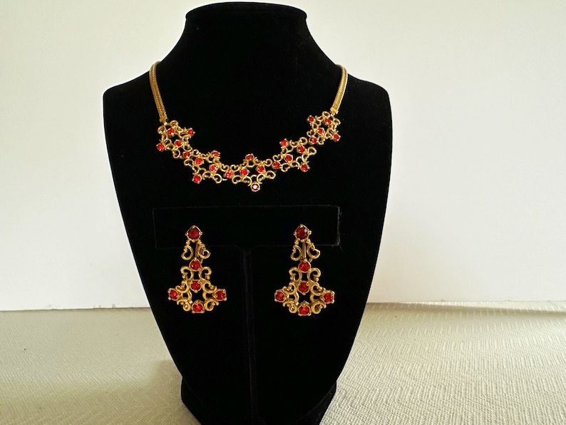 May include: A gold necklace and earrings set with red rhinestone accents. The necklace is a delicate chain with a floral design and the earrings are dangling with a similar floral design.