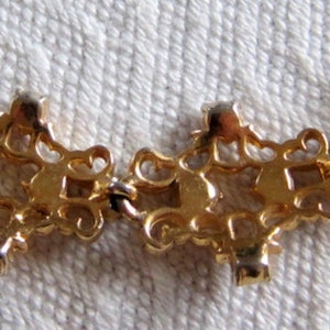 May include: Gold tone necklace with a delicate chain and a series of ornate, interlocking, pendant-like charms. Each charm features a clear rhinestone in the center.