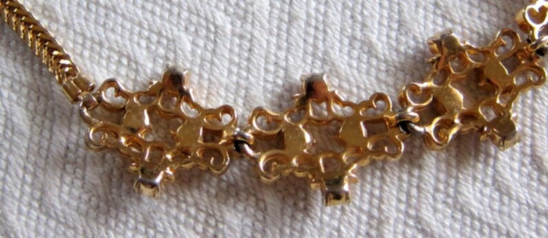 May include: Gold tone necklace with a delicate chain and a series of ornate, interlocking, pendant-like charms. Each charm features a clear rhinestone in the center.