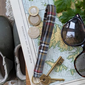 Może przedstawiać: Pasek na nadgarstek z tkaniny w kratę ze złotym brelokiem i dwoma kluczami. Pasek jest brązowy, czerwony i biały. Klucze leżą obok pary okularów przeciwsłonecznych i mapy. Widocznych jest również kilka monet.