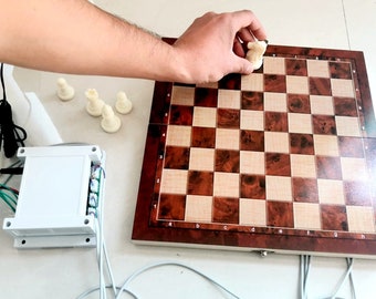 Puede incluir: Un tablero de ajedrez de madera con un patrón marrón y beige. Una pieza de ajedrez blanca está colocada en el tablero. El tablero está conectado a una caja blanca con cables. También hay piezas de ajedrez blancas en la mesa.