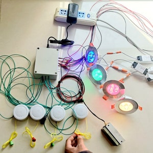 Puede incluir: Una caja blanca con cables conectados a seis botones redondos blancos, cuatro luces redondas blancas y cuatro luces redondas con LED de diferentes colores: azul, verde, rojo y blanco. Las luces están conectadas a una regleta blanca con un adaptador de corriente negro.