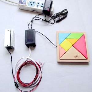 May include: A wooden puzzle with a square frame and colourful geometric pieces. The puzzle is connected to a black box with wires and a power adapter.