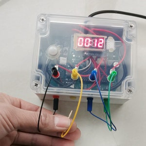 May include: A clear plastic box with a red digital display showing 00:12. The box has a white button and four wires connected to it. The wires are black, yellow, blue, and green.
