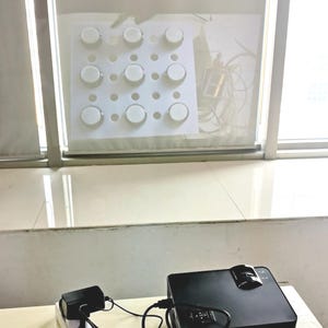 Può includere: Un proiettore nero con obiettivo e pannello di controllo è su un tavolo. Un piccolo dispositivo nero è collegato al proiettore. Sullo sfondo è visibile un pannello bianco con nove oggetti circolari.