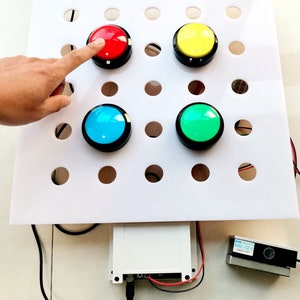 May include: A white board with a grid of holes, four large round buttons in different colours: red, yellow, blue, and green. The buttons are mounted on the board and can be pressed. A hand is pressing the red button. There is a small electronic device and a black cable connected to the board.