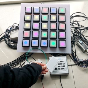 Pode incluir: Um painel branco com uma grade de 36 botões quadrados pretos, cada um com uma luz de cor diferente dentro. Uma pessoa está segurando um cartão branco com o texto "KOIBN ACCESS CARD 011111" na frente de um teclado prateado com o texto "KOIBN ACCESS CONTROL" e os números de 1 a 9, 0, *, # e A-D.