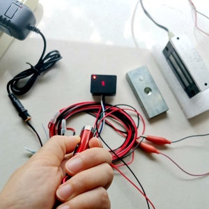 May include: A close-up of a red and black electronic device with wires and a power adapter. The device has a red light on it. The device is on a white surface.