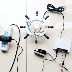 Puede incluir: Un timón de barco de estilo náutico blanco y azul con un diseño de cuerda negra, un adaptador de corriente blanco enchufado a una regleta, un dispositivo rectangular negro y gris y una caja blanca con cables conectados.