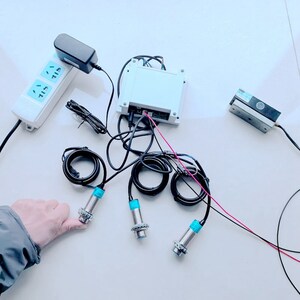 May include: A white box with wires and a power adapter connected to it. There are three blue and silver sensors with wires connected to the box. The sensors are on a white surface.