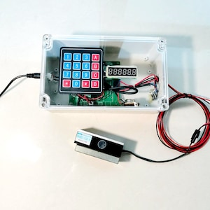 Pode incluir: Uma caixa de plástico transparente com um teclado, um visor digital mostrando "88:88:8.8" e uma placa de circuito dentro. Um sensor preto e cinza com uma etiqueta azul está conectado à caixa por um fio preto.