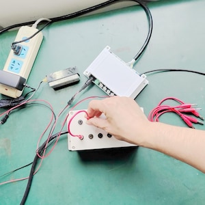 May include: A white rectangular electronic control box with four circular ports, connected to a power strip and a secondary white module via black and red electrical wires. A set of red jumper cables with metal clips is positioned to the right.