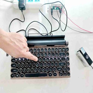 以下が含まれることがあります： 丸いキーと木製のフレームを持つヴィンテージスタイルのキーボード。キーボードは黒と銀のカラーリングで、上部にファンクションキーの列があります。キーボードはコンピューターに接続されており、手がキーを押しています。