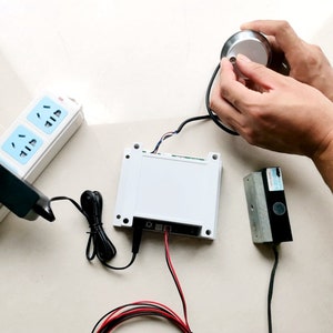 May include: A white electronic control box with a silver dial and a black power adapter. Wires connect the components, including a black and red cable. A power outlet is visible in the background.