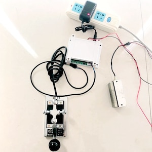 May include: A white electrical outlet with a black power adapter plugged in. A white box with a circuit board is connected to the adapter. A silver lever switch with a black handle is connected to the box. A silver magnetic lock is on the table.